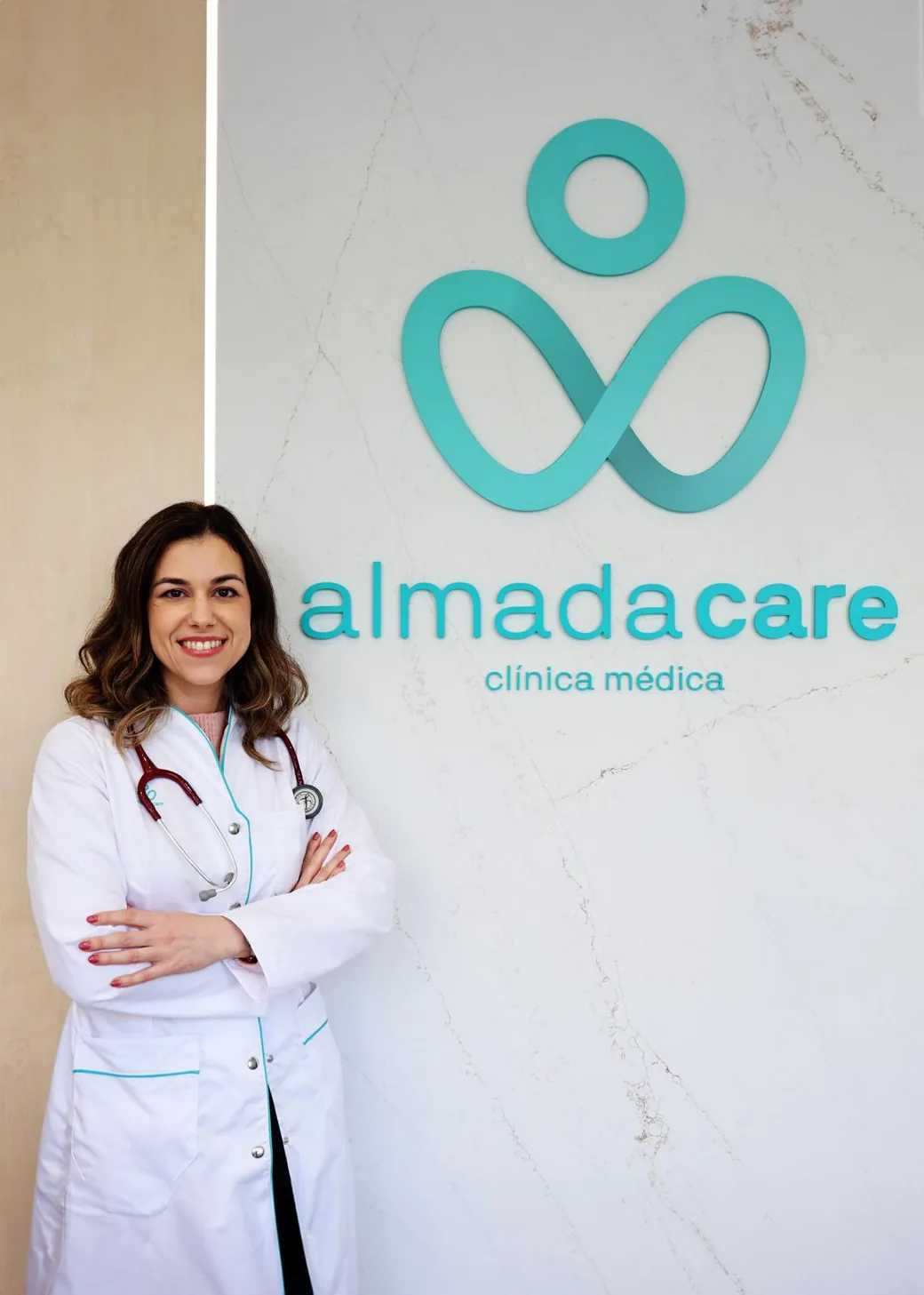 Médica da Almada Care com estetoscópio, a sorrir junto à parede da marca.